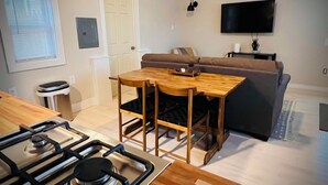 Un espacio habitable compacto con área de cocina que incluye una estufa a gas, una mesa de comedor con cuatro sillas, un sofá y un televisor de pantalla plana montado en la pared.