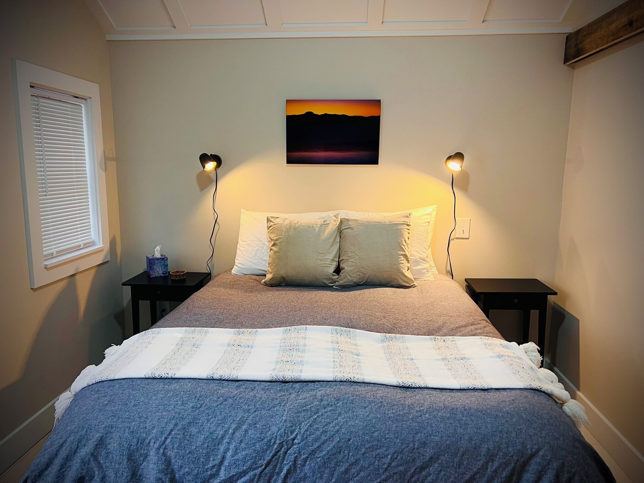 Un dormitorio con una cama, dos mesitas de noche, una mesita auxiliar, una ventana con persianas y un cuadro colgado en la pared.