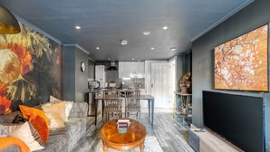 Un salon avec un canapé, une table basse et des œuvres d’art. Au fond, on aperçoit une cuisine avec des placards et un coin repas.
