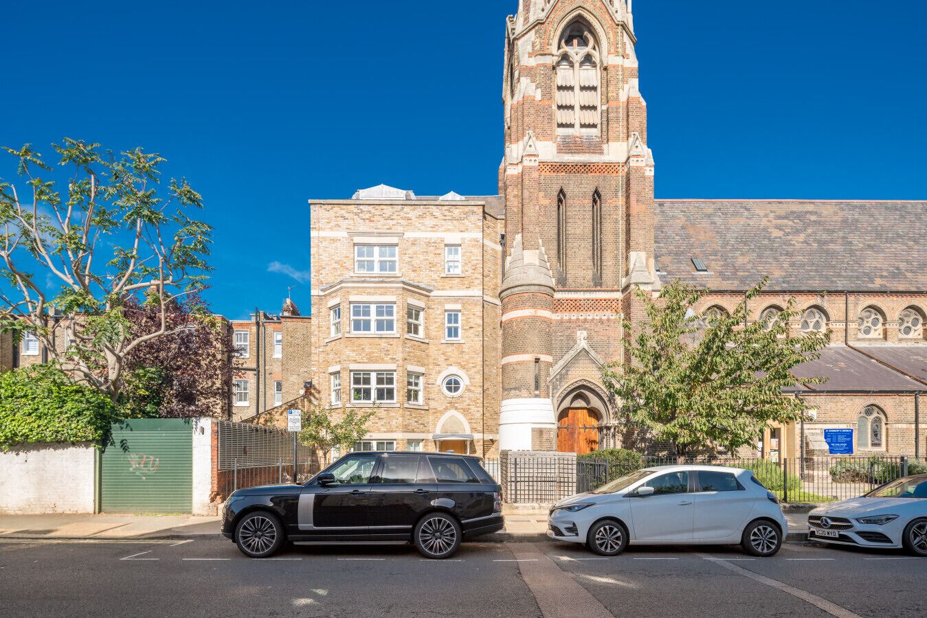A historic building with a prominent tower and arched windows, adjacent to a modern street with parked cars.