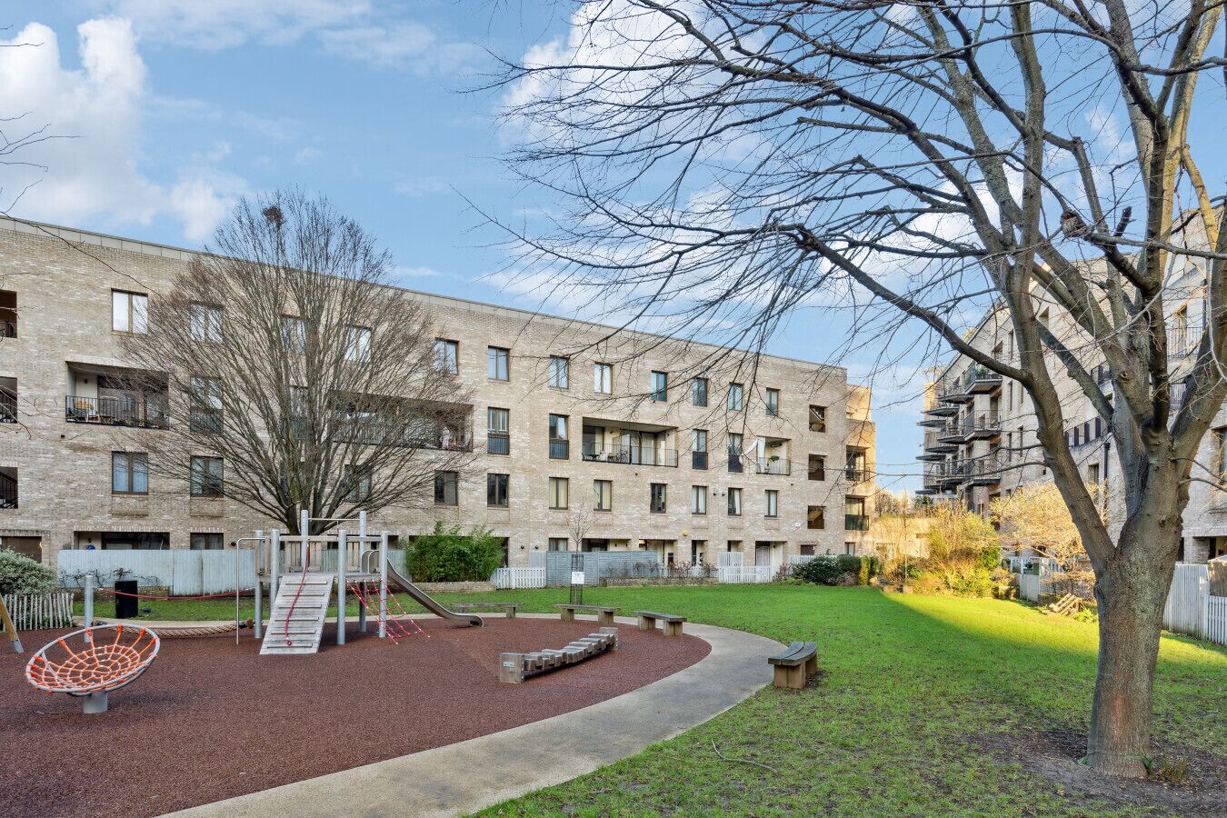 A multi-story residential building with balconies, a playground with a basketball hoop, and a tree in the foreground.