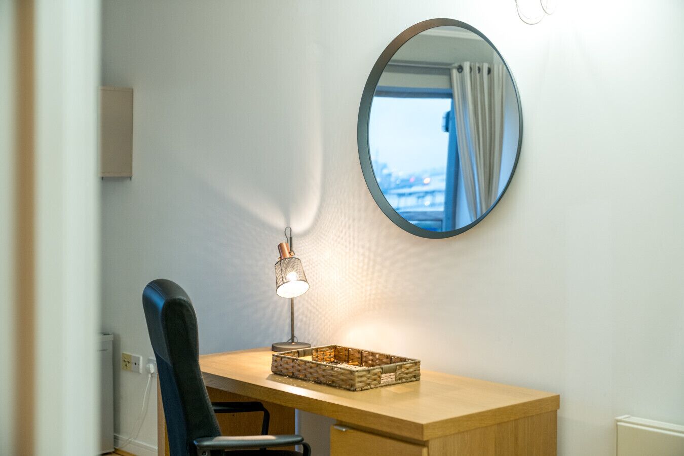 Una habitación pequeña con un escritorio de madera, una silla negra, un espejo redondo y una lámpara pequeña.