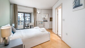 A modern hotel room with a large bed, a desk, a chair, a window with curtains, a small desk lamp, and a framed picture on the wall. - Lovely 3BD Flat, Parking, sleeps SIX close to O2 (London)