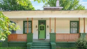 A two-story house with a green front door, a small porch with wooden railings, and a chimney on the roof.