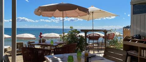 An outdoor patio with wooden tables and chairs, large umbrellas, and a view of the ocean.