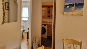 A hallway leading to a kitchen with a washing machine, a chair, and a painting of boats on water. - Beautiful, comfortable vacation apartment for 4 people in Cavalaire sur Mer. (Cavalaire-sur-Mer)