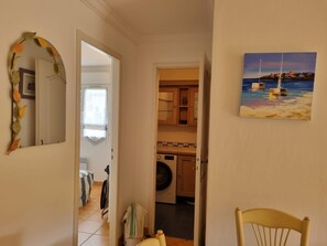 A hallway leading to a kitchen with a washing machine, a chair, and a painting of boats on water. - Beautiful, comfortable vacation apartment for 4 people in Cavalaire sur Mer. (Cavalaire-sur-Mer)