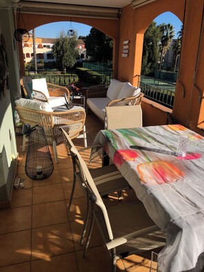 Een balkon met rieten meubels, een tafel met een kleurrijk tafelkleed, en een uitzicht op een gebouw en bomen in de verte.