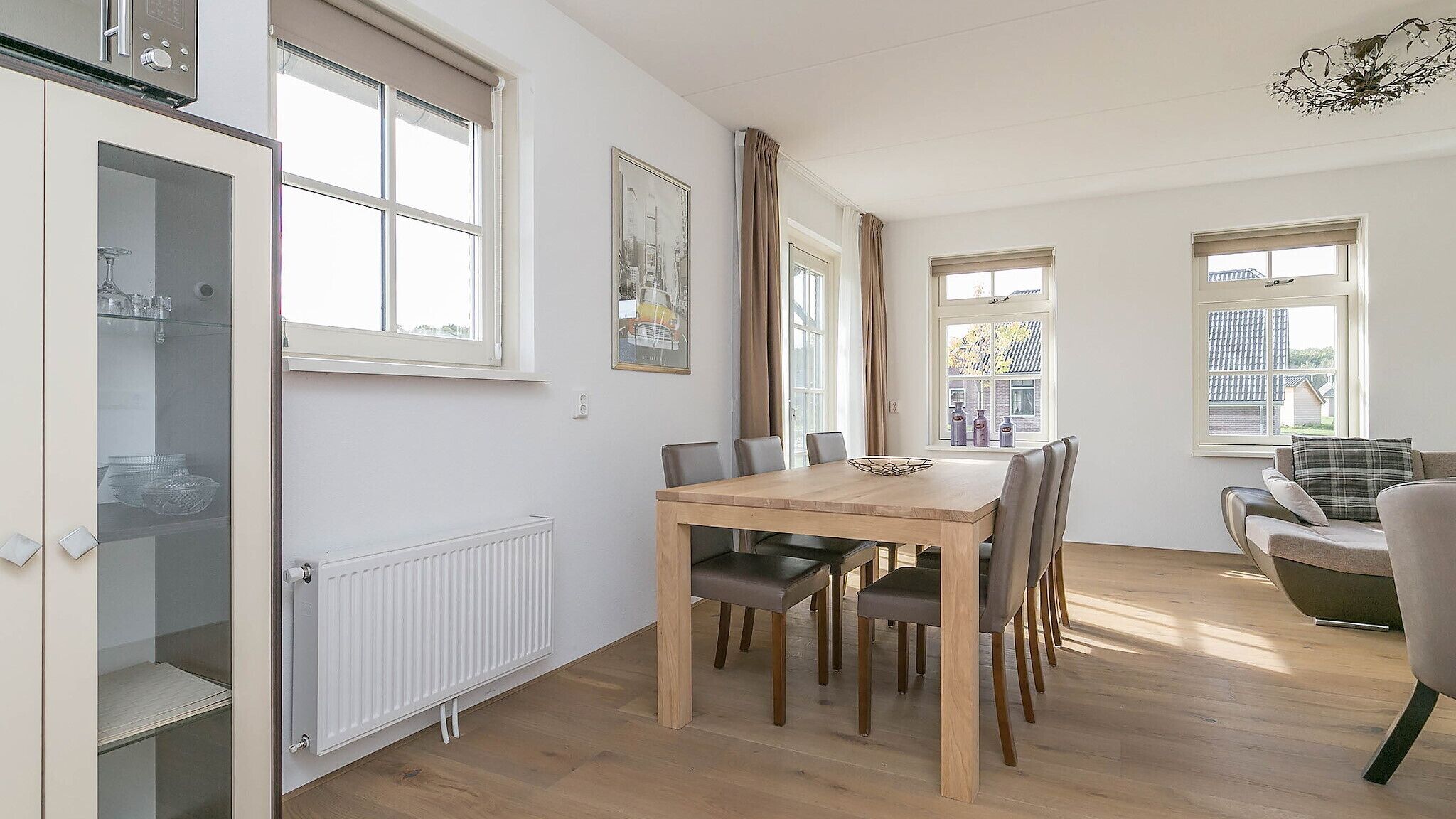 A dining area with a wooden table and chairs, a radiator, and windows with curtains.