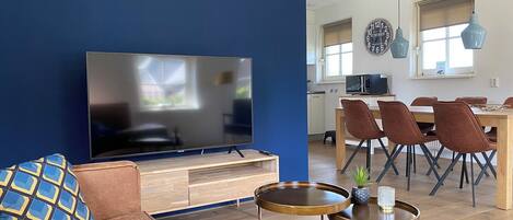 Uma sala de estar com uma televisão de tela plana grande, um sofá, uma mesa de centro e um espaço de refeições com cadeiras e uma mesa.