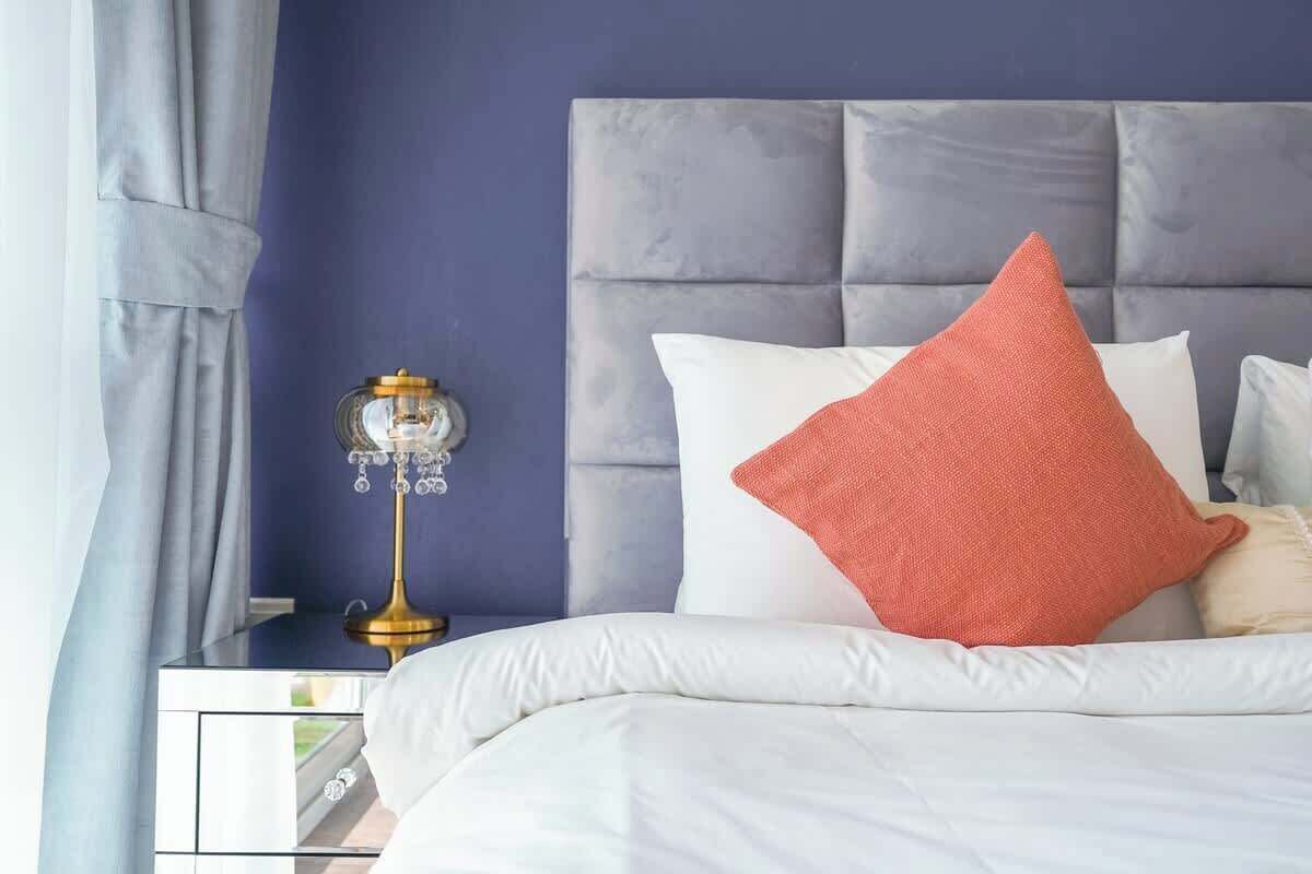 A neatly made bed with white bedding and a single orange pillow, a bedside table with a lamp, and a dark blue wall.
