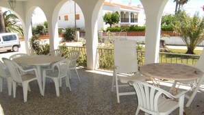Un patio al aire libre con sillas y mesas de plástico blanco, un automóvil estacionado afuera y un edificio con techo rojo al fondo.