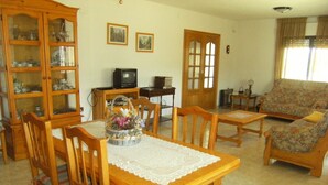 Una sala de estar acogedora con una mesa de comedor de madera, sillas, un centro de mesa floral, un televisor y un sofá.
