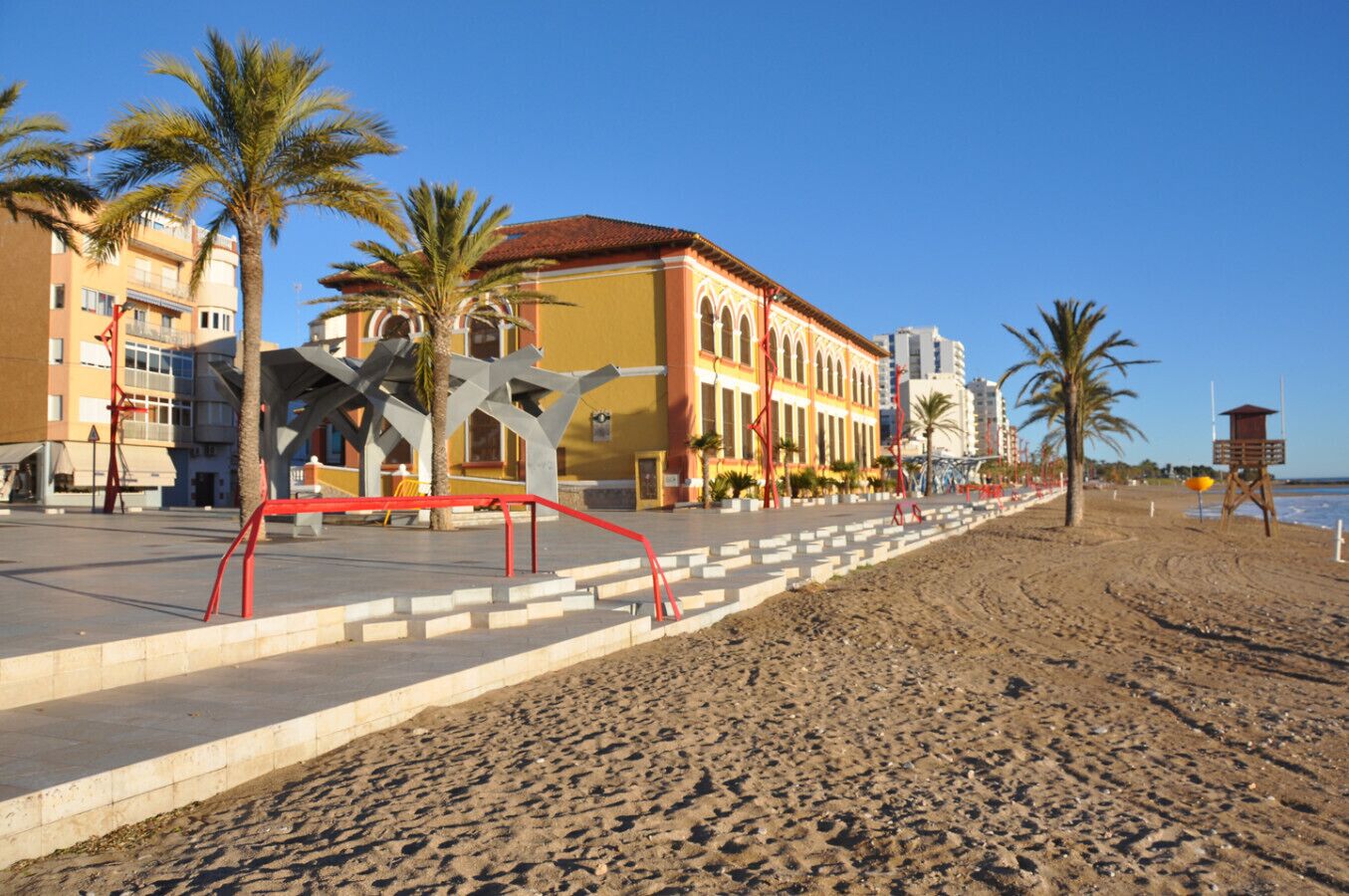 A beachside promenade with palm trees, a red metal sculpture, and buildings with balconies.