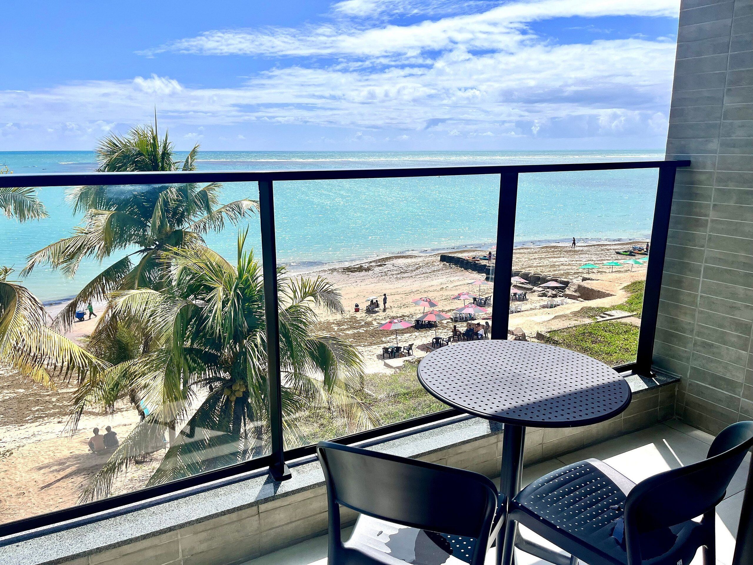 A balcony with a view of a beach, palm trees, and a table with chairs.
