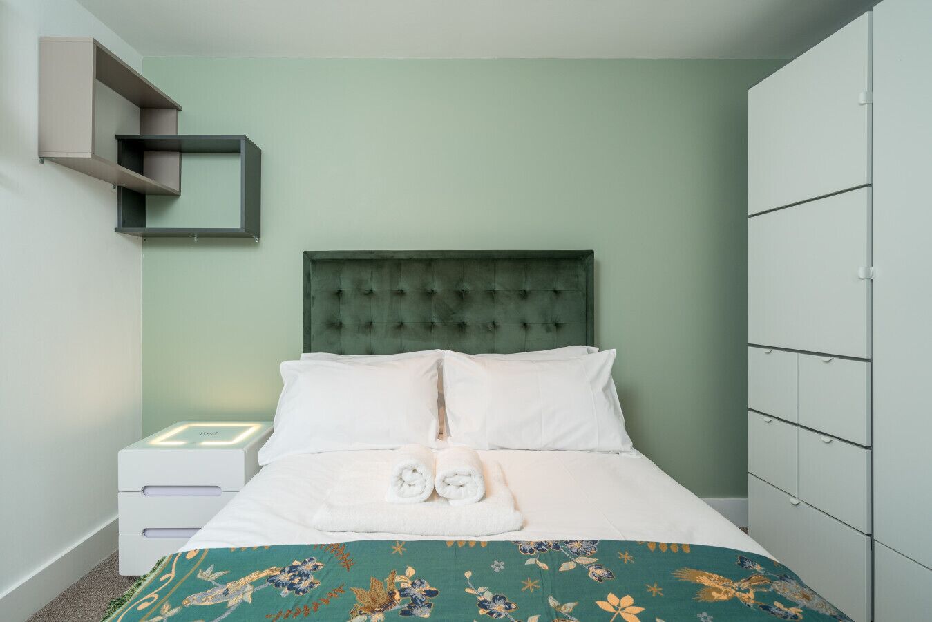 A neatly made bed with white linens and a floral bedspread, a green upholstered headboard, and a white bedside table with a lamp.