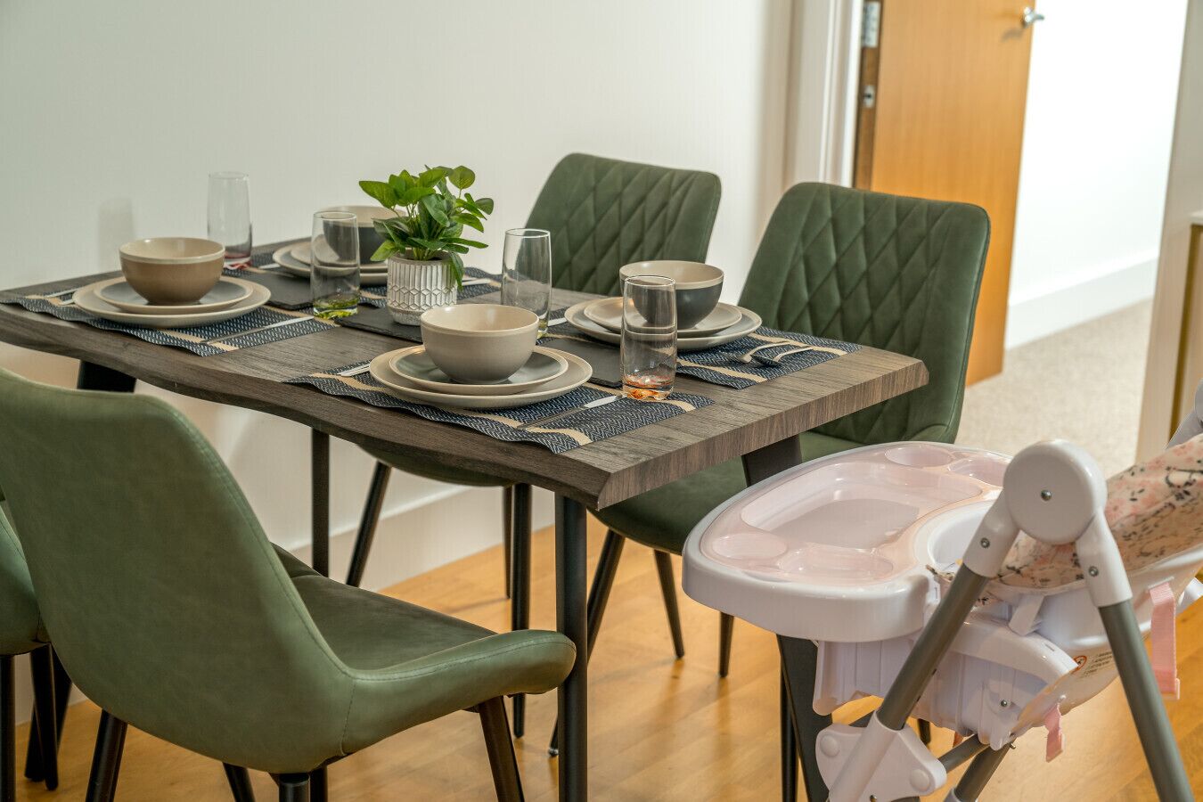A dining table set with plates, bowls, glasses, and a potted plant. There are green upholstered chairs and a high chair with a tray.