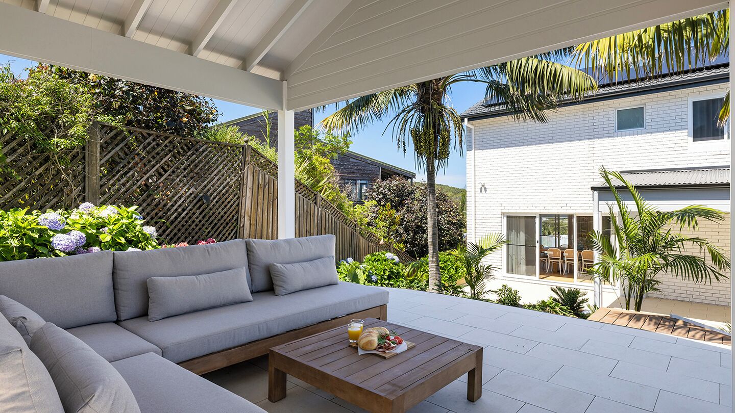 Un espace de vie extérieur moderne, avec un canapé gris, une table basse en bois et une vue sur un bâtiment blanc doté d’un balcon.