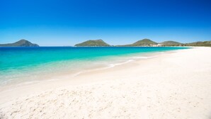 En strand med kristallklart turkost vatten, vit sand och gröna kullar i bakgrunden.