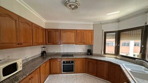 A kitchen with wooden cabinets, a microwave, an oven, granite countertops, and a window with blinds.