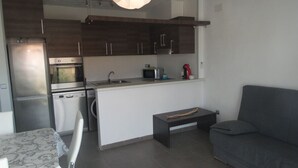 Een moderne keuken met donkere houtkasten, roestvrijstalen apparatuur en een wit aanrecht. In de woonruimte staat een grijze bank en een kleine tafel.