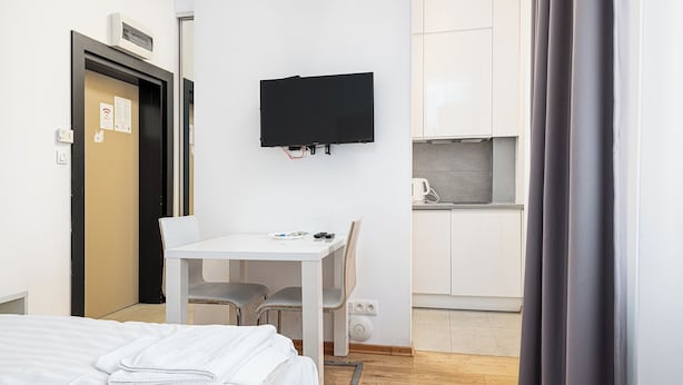 Habitación de hotel con cama, escritorio, silla, televisor montado en la pared y una kitchenette al fondo.