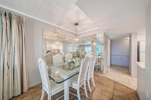Un coin repas avec une table en verre et des chaises blanches, un lustre et une vue sur la cuisine.