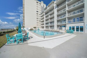 Un espace piscine doté de chaises longues et d’une table, entouré d’un bâtiment blanc comportant de nombreux balcons.