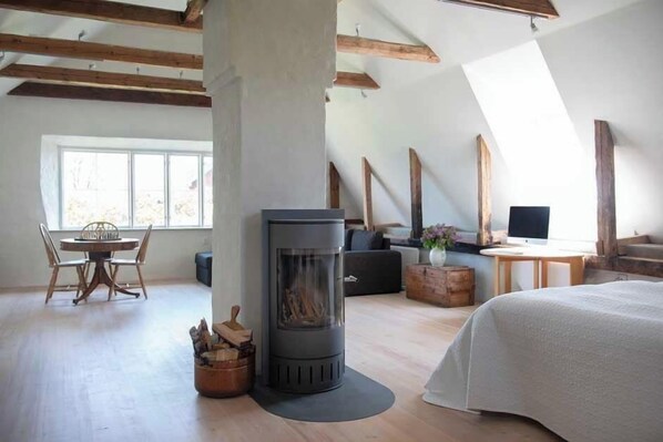 A modern loft bedroom with a central wood-burning stove, a dining area, and a large window providing natural light.
