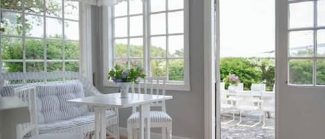 A room with a white wicker sofa, a white table, and chairs. There are large windows with white curtains.
