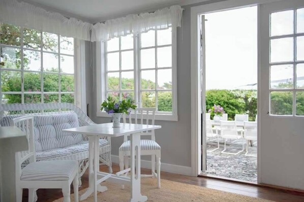 A room with a white wicker sofa, a white table, and chairs. There are large windows with white curtains.