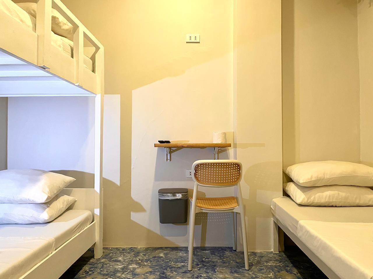 Una habitación pequeña y funcional con literas, una silla, un estante con una toalla y un bote de basura.