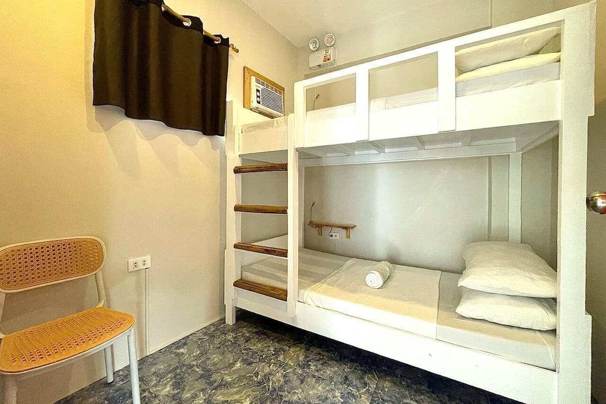 Ein Stockbett mit Holzleiter, einer kleinen Ablage und einem weißen Kissen auf der unteren Liege. Ein Korbstuhl und ein Fenster mit Vorhang sind ebenfalls vorhanden.