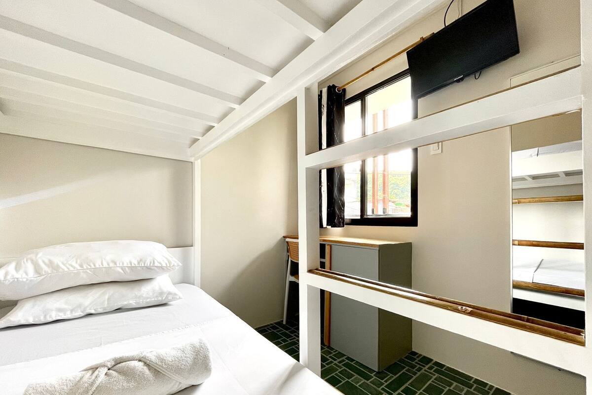 Ein kleines Hotelzimmer mit einem Stockbett, einem Schreibtisch mit Stuhl, einem Fernseher und einem Fenster mit Vorhängen.