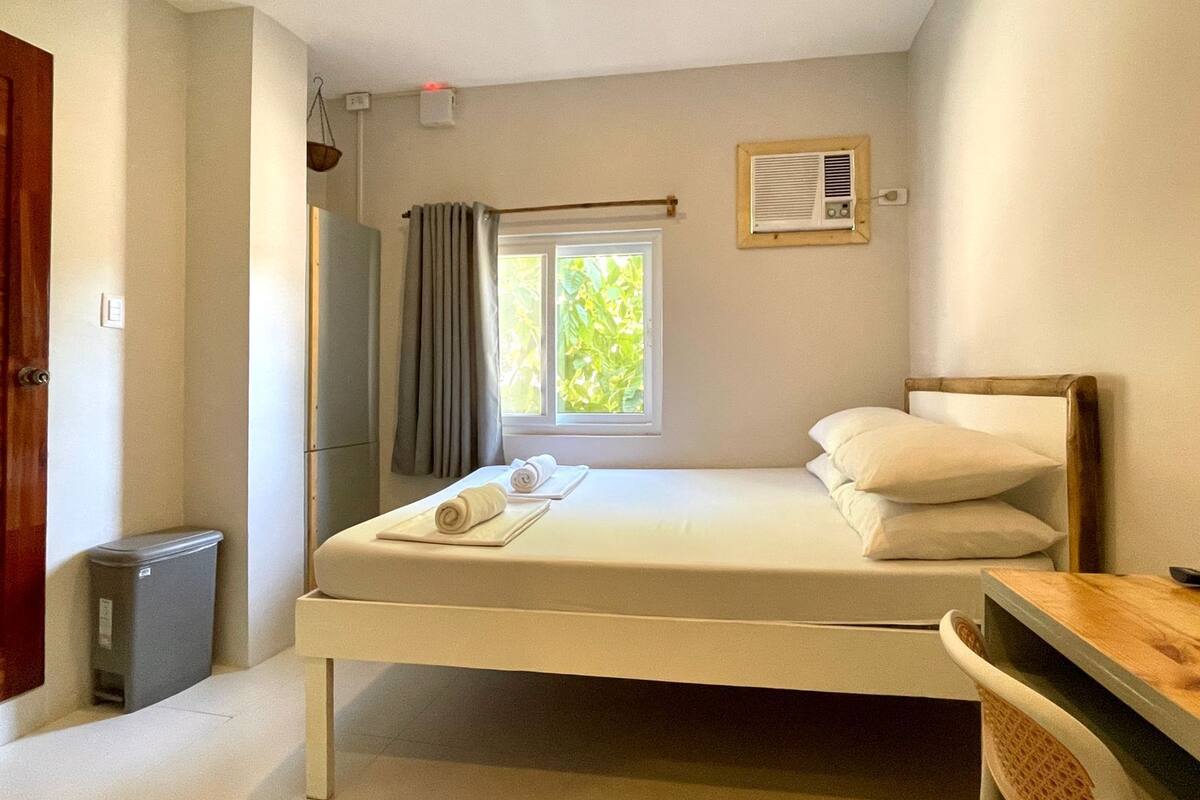 Ein Schlafzimmer mit einem Bett, einem Fenster mit Vorhängen, einem Heizkörper und einer kleinen Mülltonne.