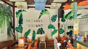 Interno di un ristorante con un murale che pubblicizza un evento karaoke, tavoli e sedie in legno e un bancone con una ventola e opzioni di pagamento.
