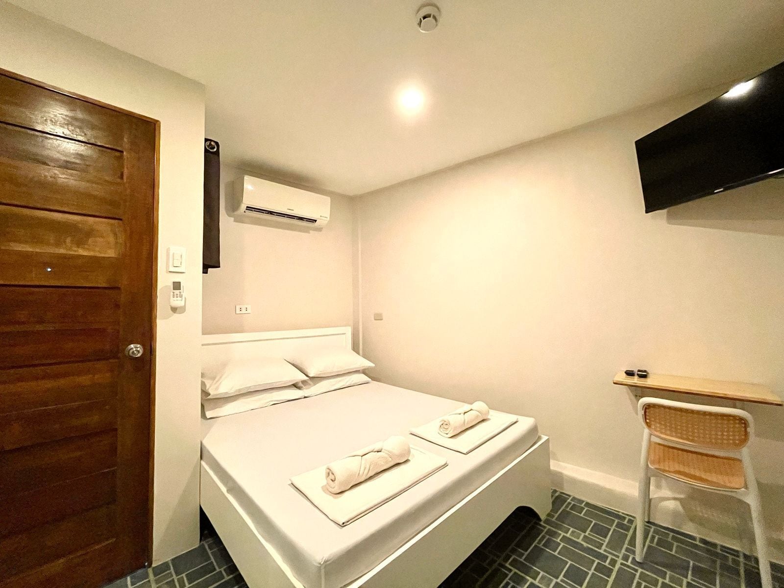 A bedroom with a bed, a wooden door, an air conditioning unit, a TV mounted on the wall, and a small table with a chair.