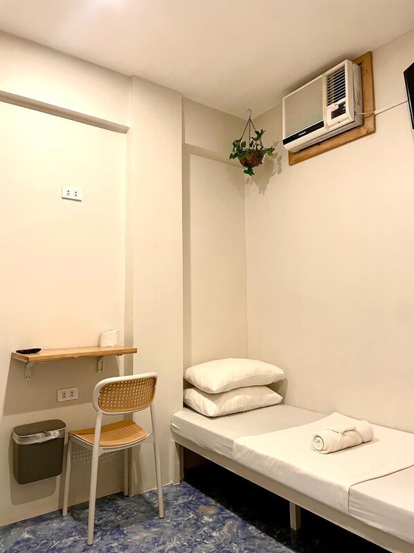 Une petite pièce propre, avec un lit simple, une chaise, une petite étagère et un appareil de climatisation.
