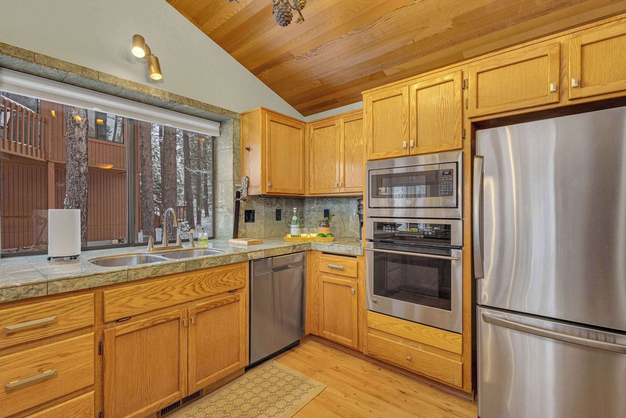 A modern kitchen with wooden cabinets, stainless steel appliances, and a large window overlooking a wooden deck.