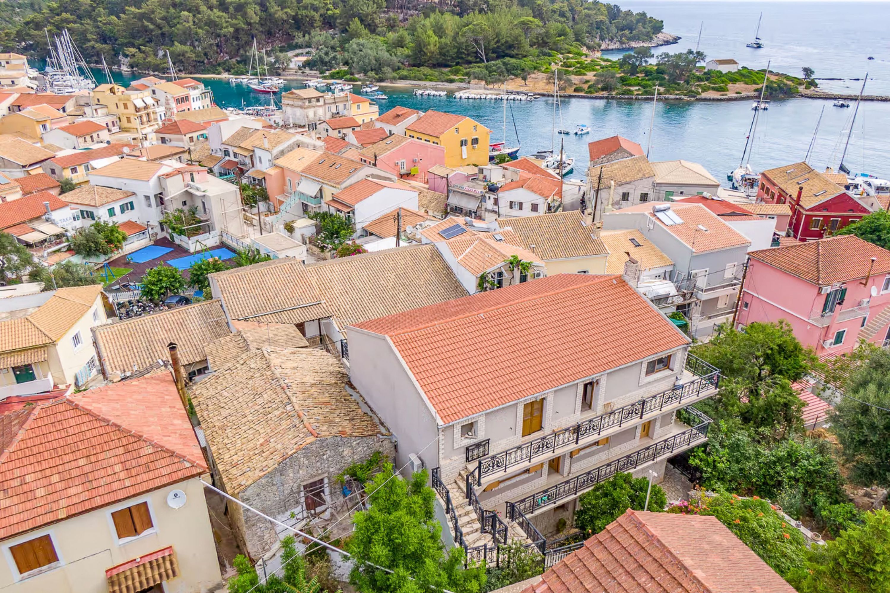 Un pueblo costero con edificios de tejados de terracota, un puerto deportivo con barcos y una ladera cubierta de vegetación.