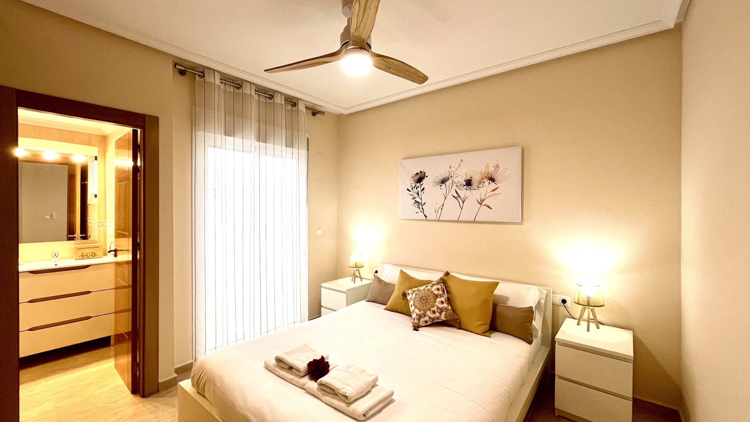 A hotel room with a bed, bedside tables, a ceiling fan, a mirror, and a floral artwork on the wall.