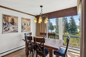 A dining area with a wooden table and chairs, two posters on the wall, and a view of trees through large windows. - Stunning 2BR on Lake Dillon w/ Pool and Concert Access! (Dillon)