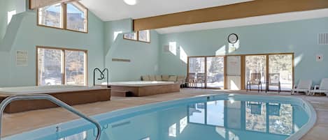 An indoor swimming pool with clear glass walls, a lounge area with chairs, and a view of the outside.