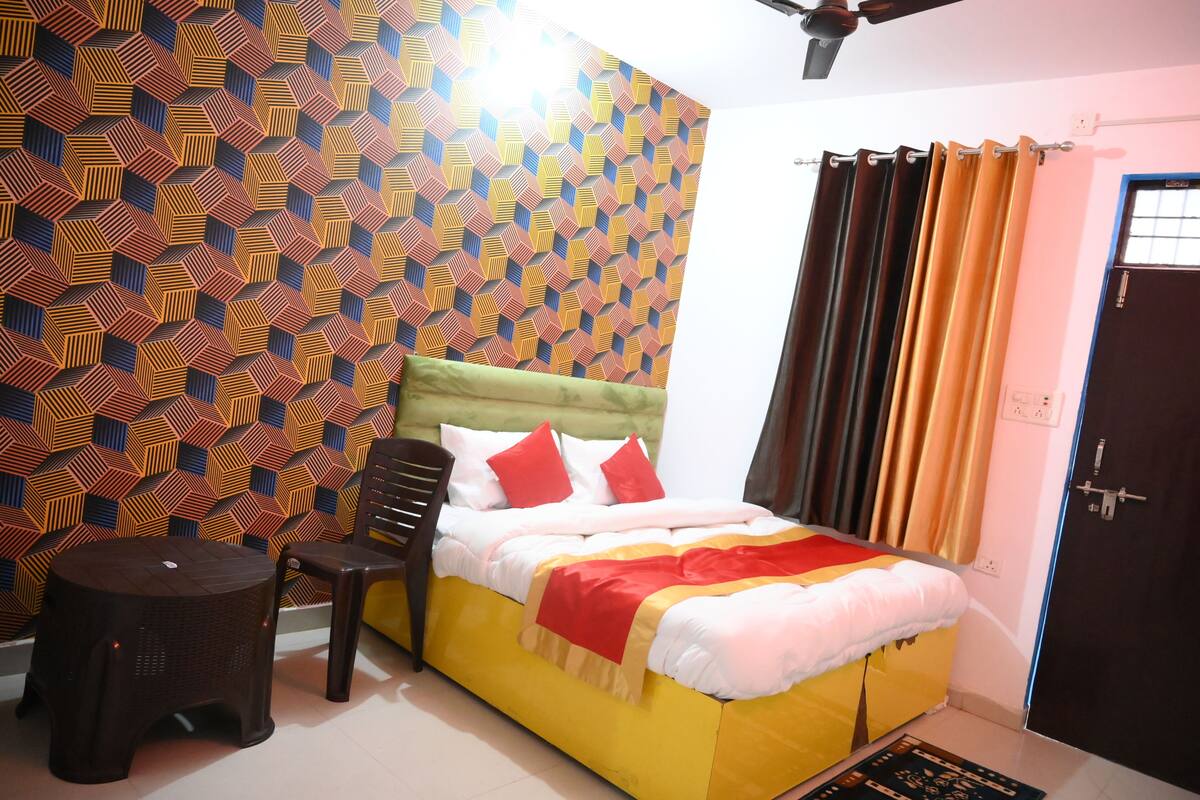 A room with a patterned wall, a bed with a yellow and white bedspread, a green headboard, and a window with curtains.