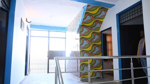 A hallway with a metal railing, a colorful wall with a wavy pattern, and a large window allowing natural light. - All In One Guest House (Bareilly)