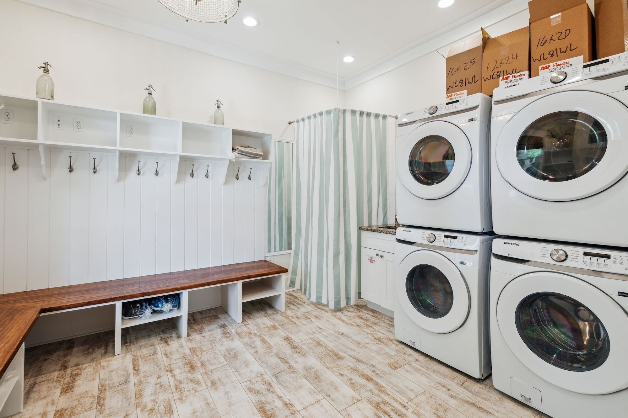A laundry room with multiple washing machines, a bench, shelves with bottles, and striped curtains.