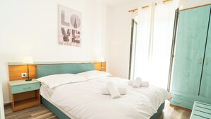 Een net opgemaakt bed met witte lakens en handdoeken, een houten nachtkastje met een lamp, een blauwgroene garderobe en een poster van een stadsgezicht aan de muur.