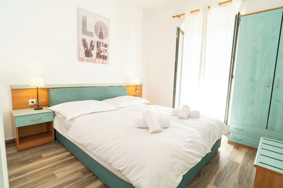A neatly made bed with white linens and towels, a wooden nightstand with a lamp, a teal-colored wardrobe, and a cityscape poster on the wall.