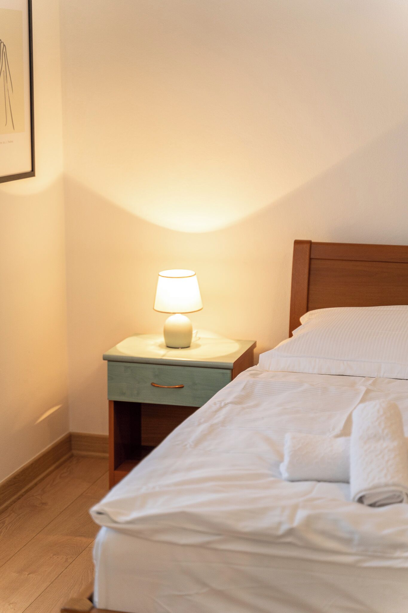 Una cama bien hecha con sábanas blancas, una lámpara de noche en una mesita y un cuadro en la pared.