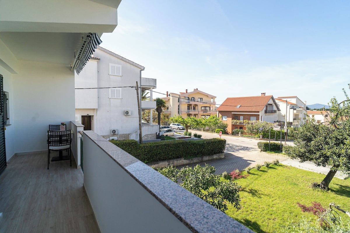 A balcony with a chair and a small table, a garden with hedges and a tree, and a street with cars and buildings in the background.